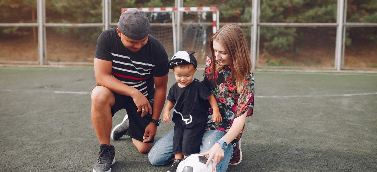 Full body of positive smiling family with cute little boy resting on football field with ball against football gate
