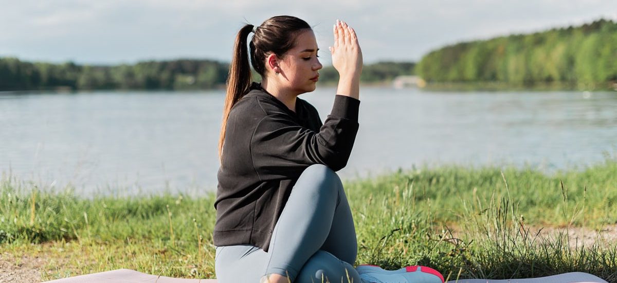 Young woman meditating outdoors on a yoga mat by the lake, enjoying a peaceful moment.